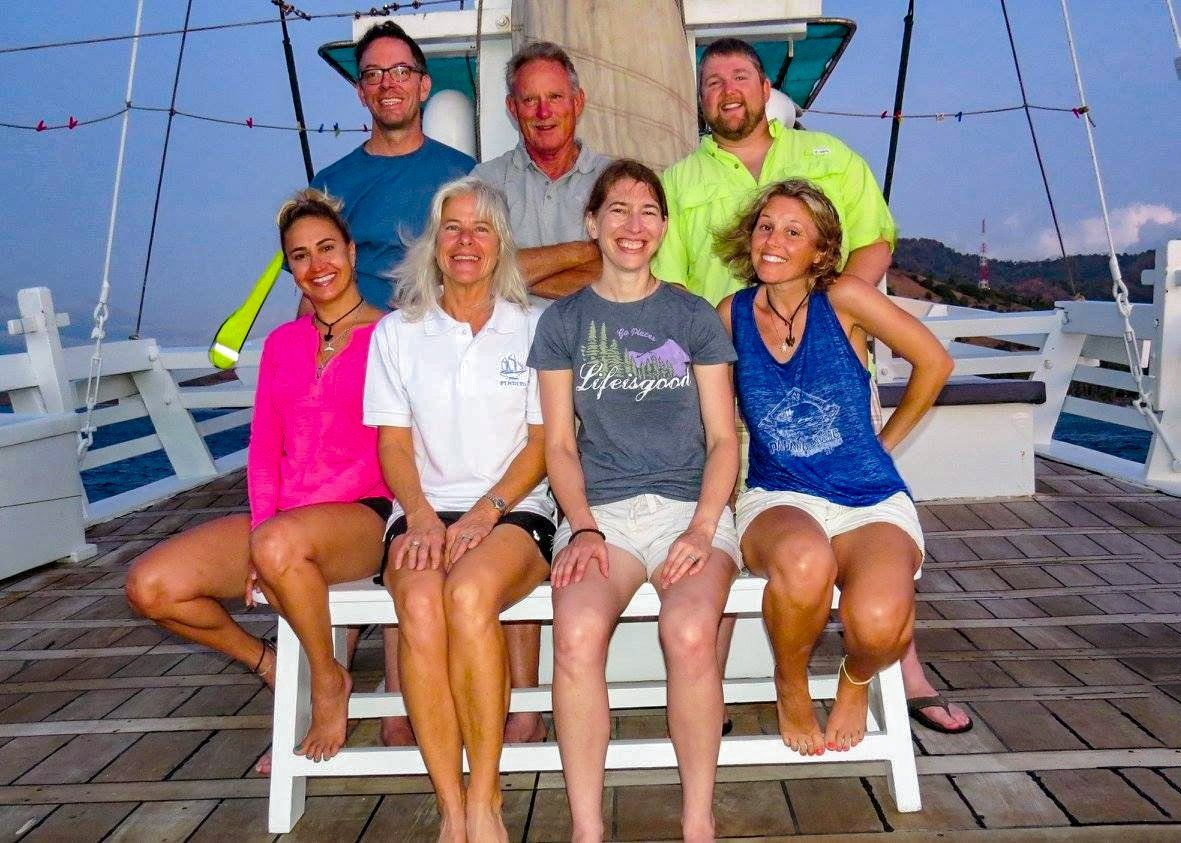 Group of people on a boat deck smiling. Sitting and standing, mostly wearing casual clothes. Evening setting.