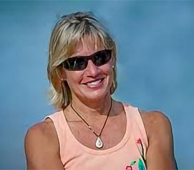 Woman wearing sunglasses smiles outdoors. Light orange top, pendant necklace. Blue background.