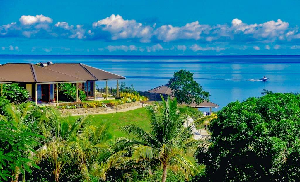 Lush green tropical landscape with a building overlooking a calm blue ocean, under a partly cloudy sky.