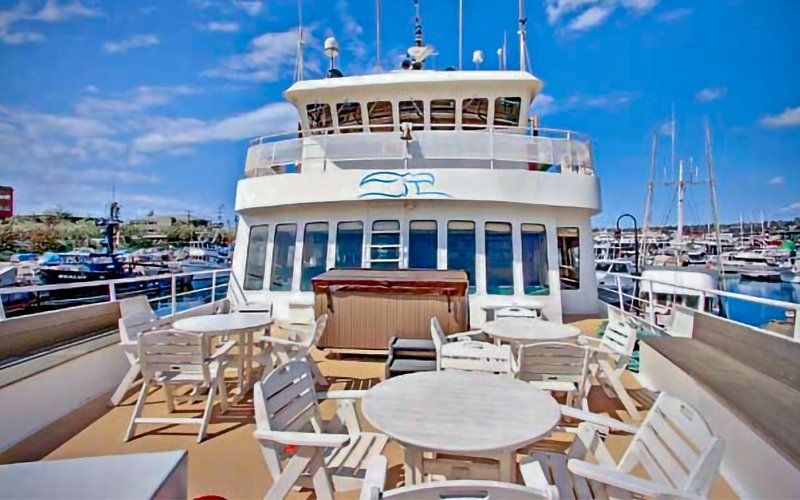 Back deck of a white boat with tables, chairs, and a hot tub overlooking a harbor on a sunny day.