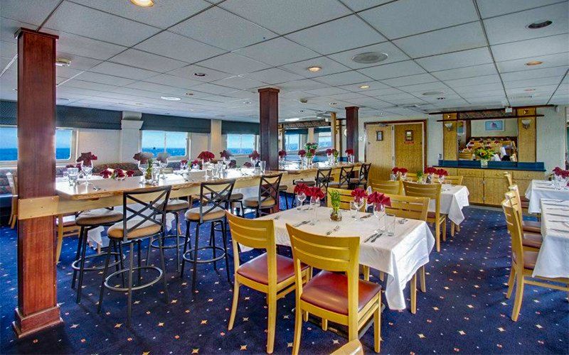 Restaurant interior with tables set for dining, red flower centerpieces, blue carpet, and ocean view windows.