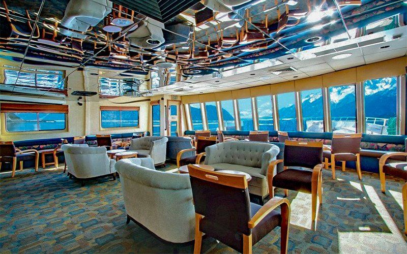 Lounge with large windows showcasing mountain and water views; seating includes sofas and chairs. Mirrored ceiling.