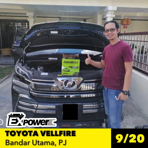 Man standing beside black Toyota Vellfire with open hood, holding a key; storefront backdrop and yellow plate displayed