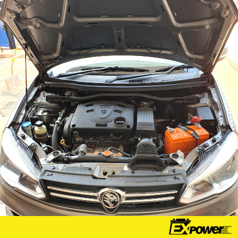 Car engine bay with hood open, showing an orange battery and black engine components.