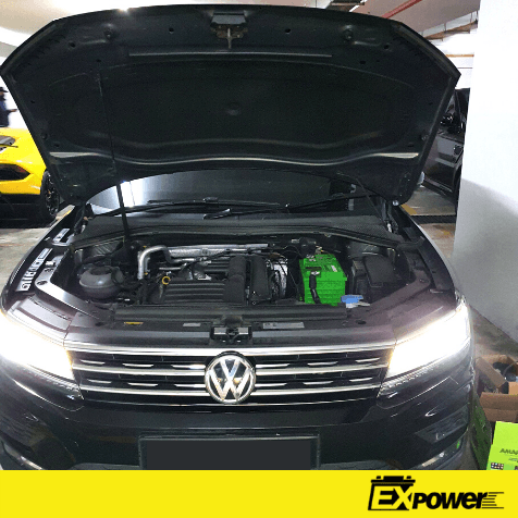 Open car hood showing engine bay, with bright green battery and Volkswagen grille in a garage