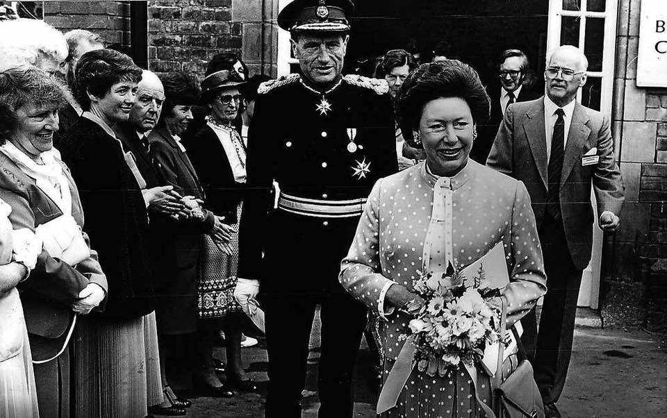 Princess Margaret holding flowers, followed by uniformed man and others, outside building.