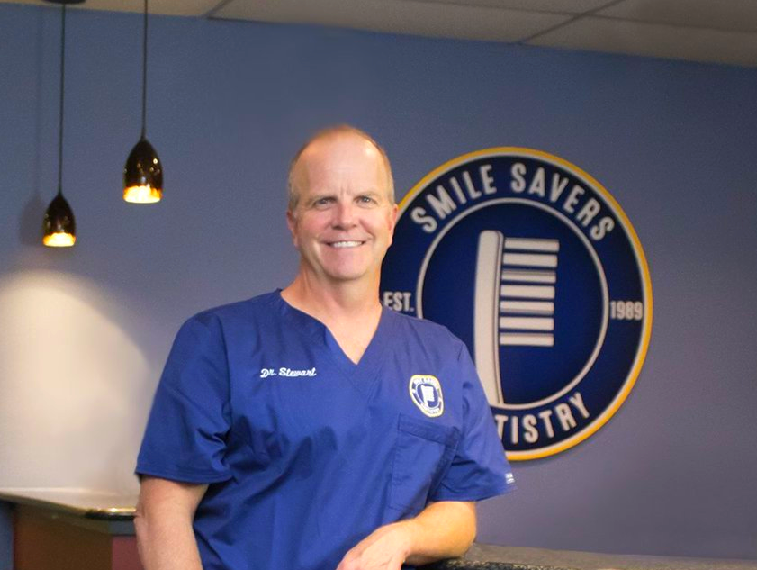 Man in blue scrubs at Smile Savers Dentistry. Smiling in front of a dental office logo.