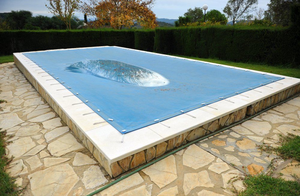 Rectangular pool covered with blue tarp, surrounded by stone patio and hedges.