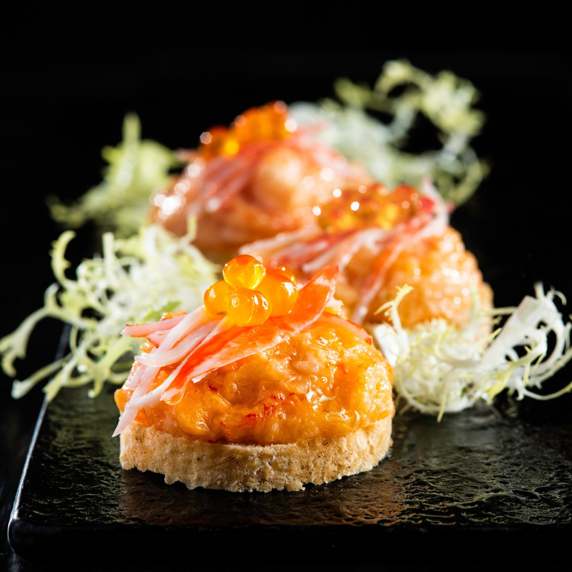 Sushi bites with orange filling and roe, topped with crab on a black plate.