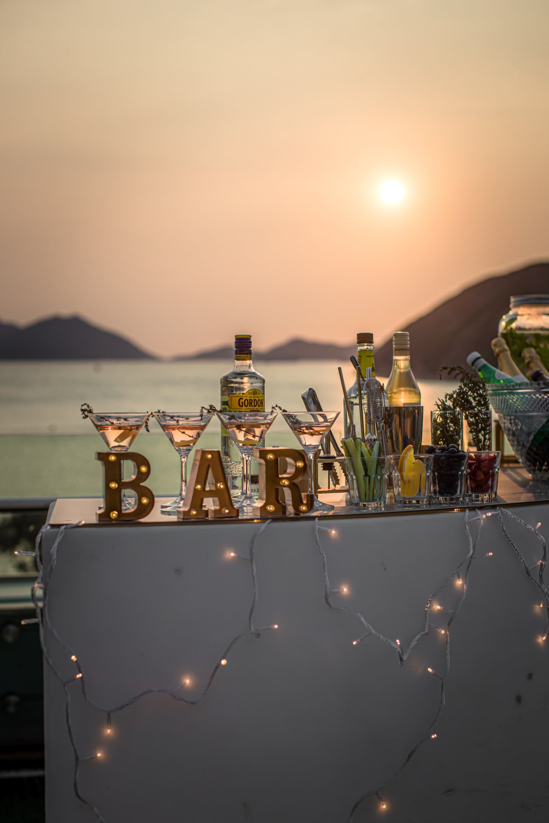 Bar setup at sunset with cocktails, bottles, and string lights.