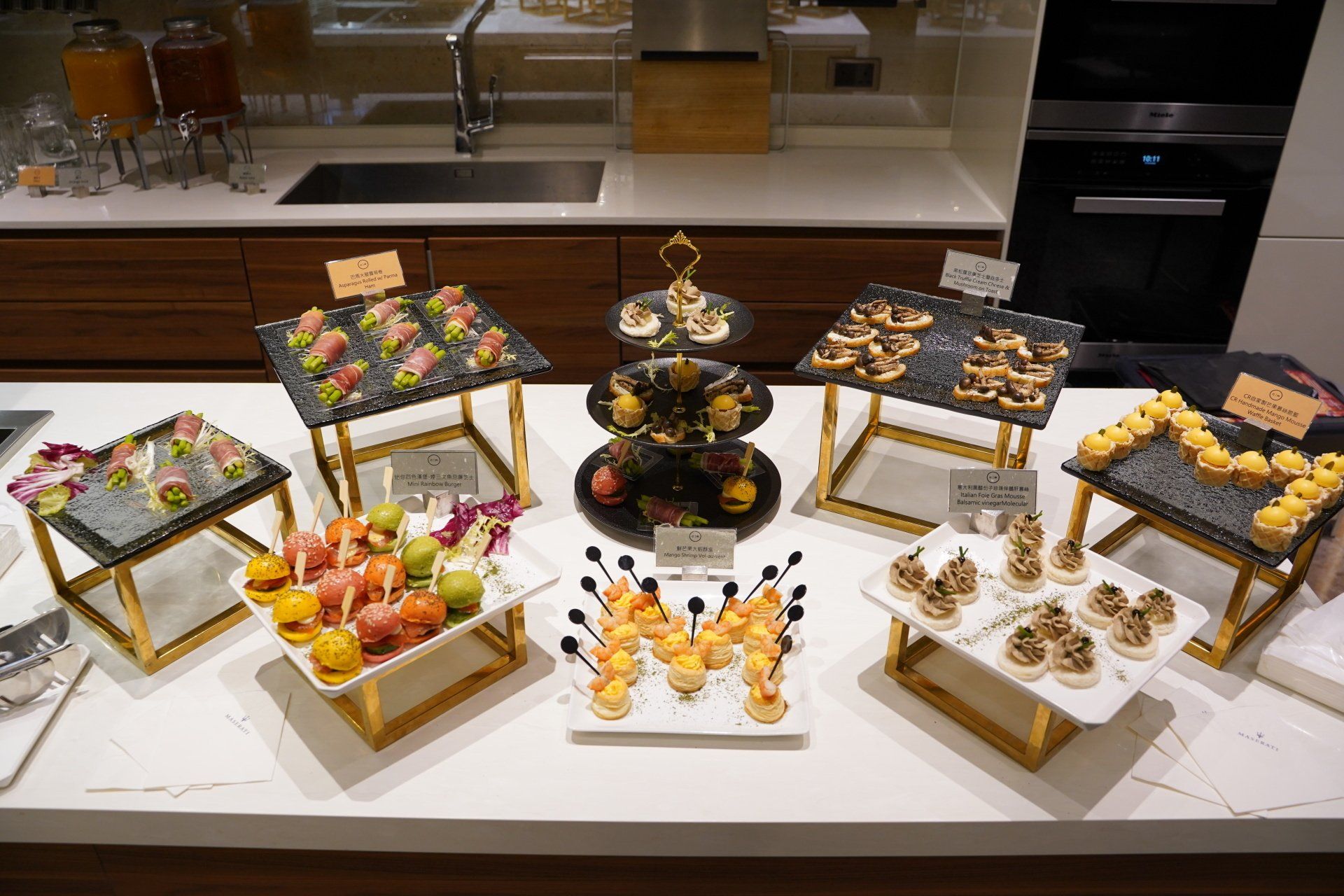 A buffet spread of appetizers on a white countertop. Various bite-sized foods are arranged on trays and stands.