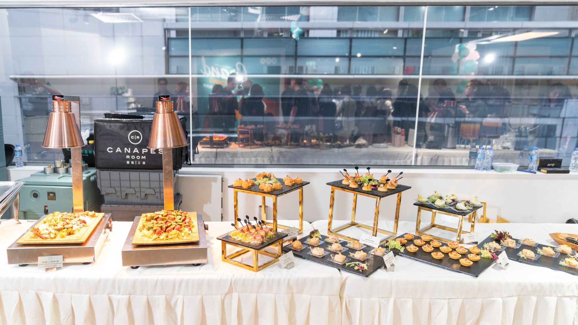 Buffet table with appetizers under copper heat lamps, in front of a glass wall.