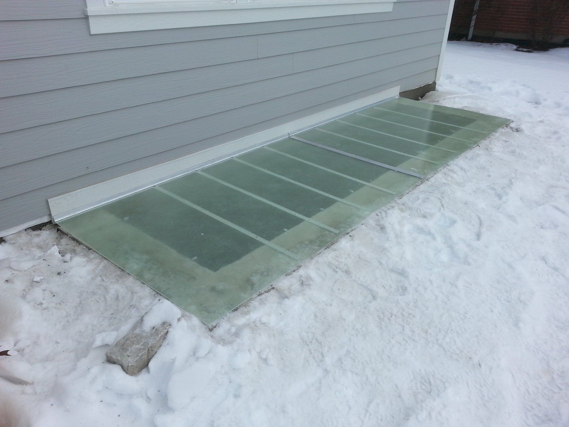 A window well cover is sitting in the snow next to a house.