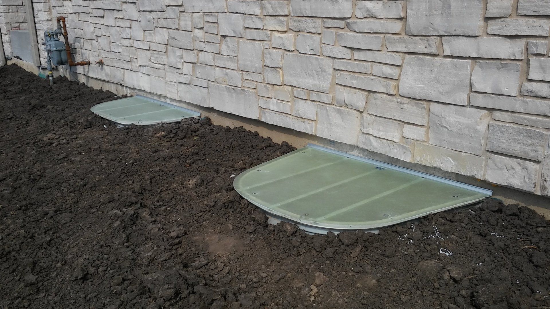 Two window wells are sitting in the dirt next to a stone wall.