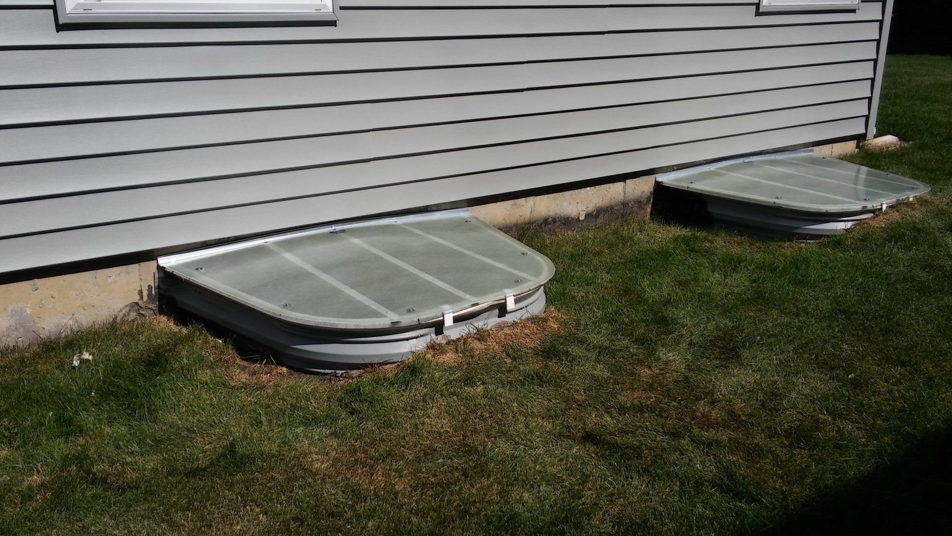 There are two window wells in the grass next to a house.
