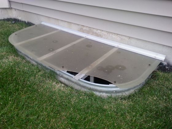 A window well cover is sitting in the grass next to a house.