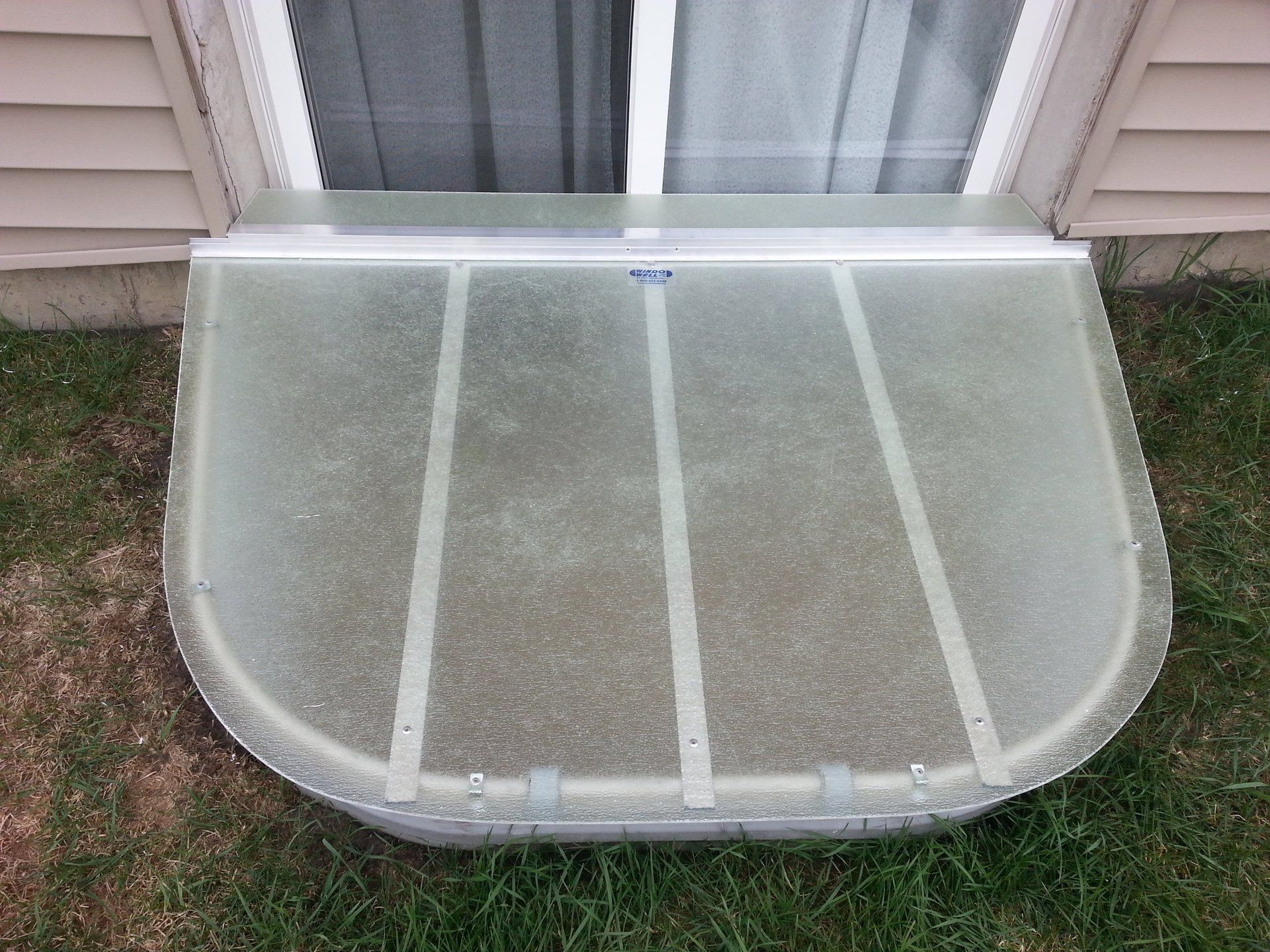 A window well cover is sitting in the grass in front of a house.