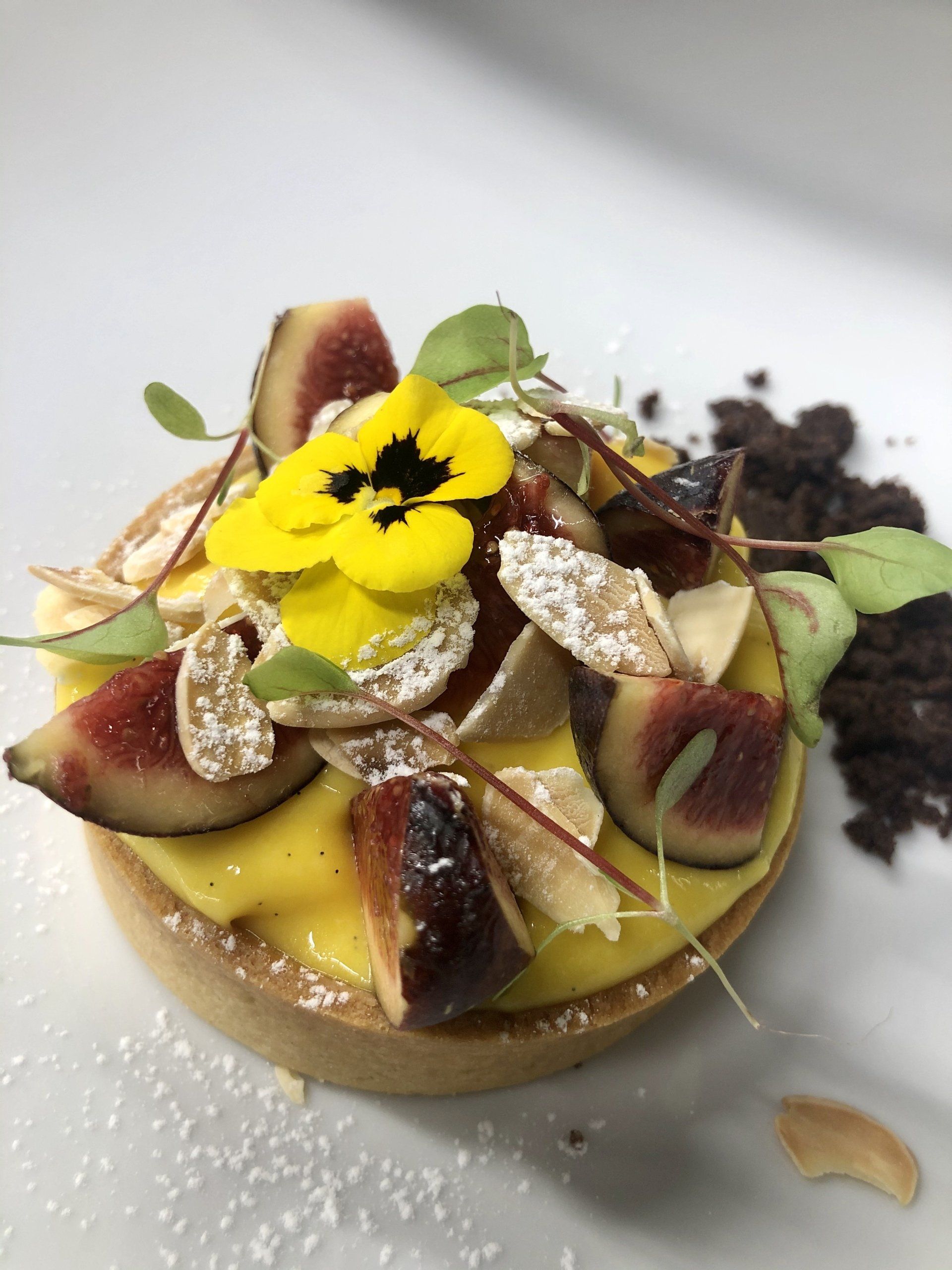 Torta dolce guarnita con fette di fichi, fiori gialli e zucchero a velo su un piatto bianco.
