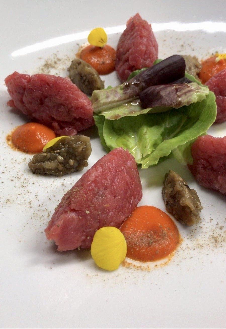 Piatto gourmet impiattato con pezzi di carne rossa, lattuga, gocce di salsa e guarnizioni su un piatto bianco.