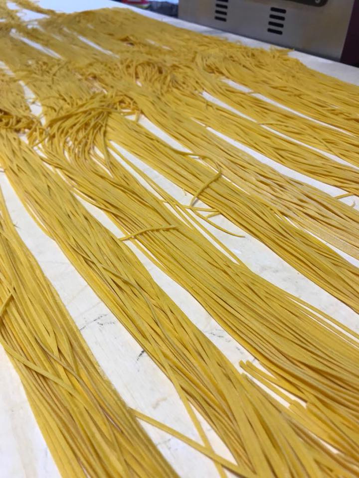 Fili di pasta gialla che si asciugano su vassoi bianchi in una cucina o in un'area di lavorazione alimentare.