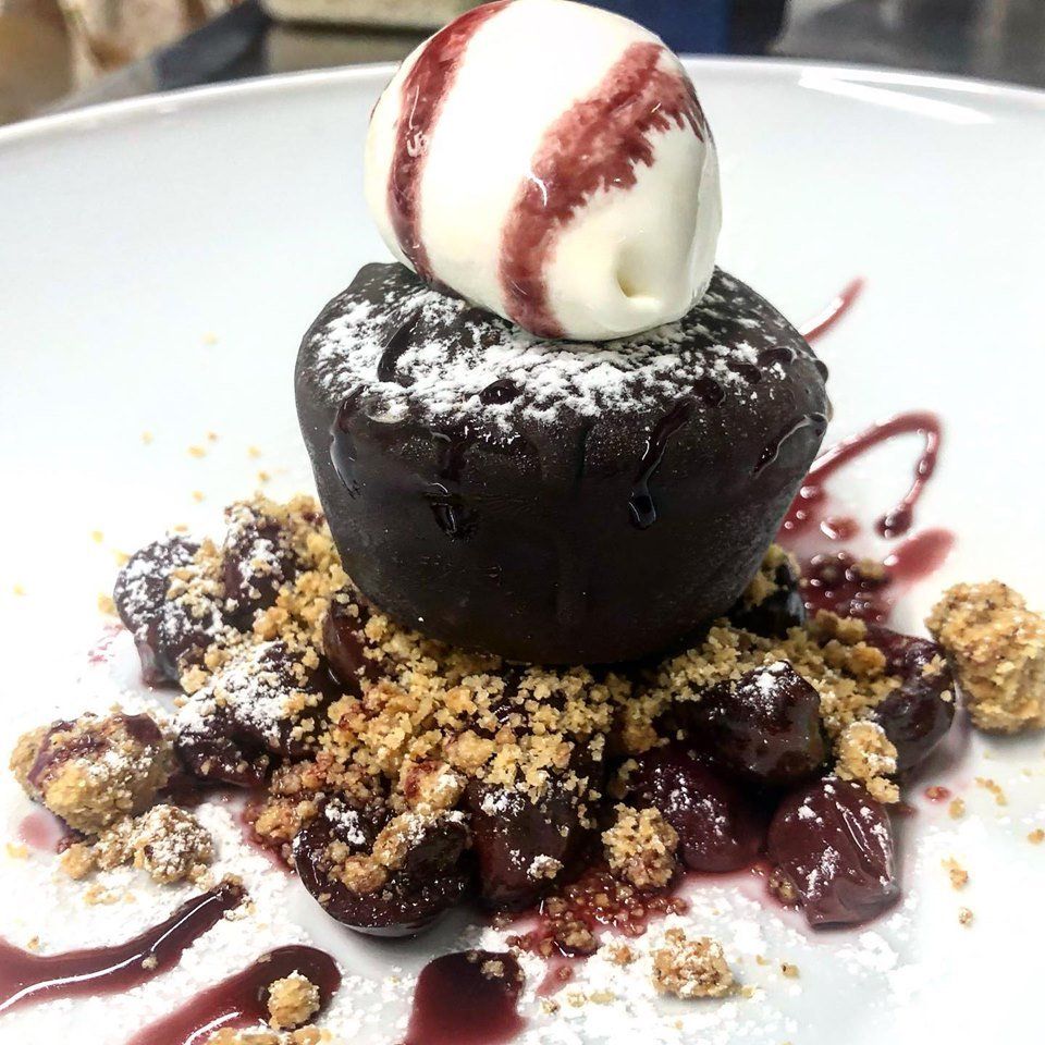 Torta al cioccolato fondente con cuore caldo, guarnita con gelato alla vaniglia e salsa ai frutti di bosco, cosparsa di briciole e servita su un piatto bianco.