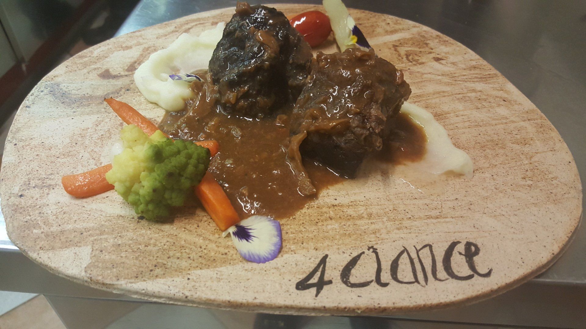 Piatto gourmet impiattato con carne in salsa bruna, verdure e salsa su un tagliere rotondo di legno.