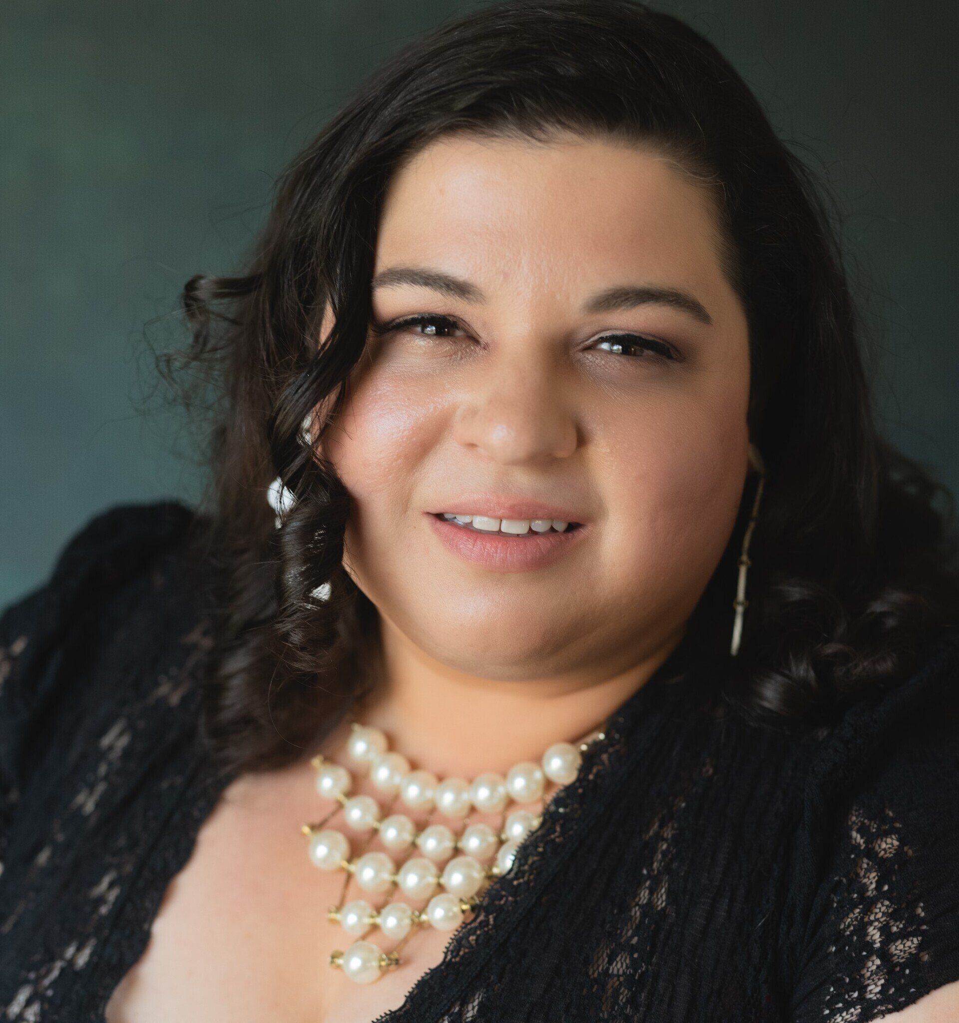 Woman with dark curly hair, wearing a pearl necklace and lace top, smiling at the camera.