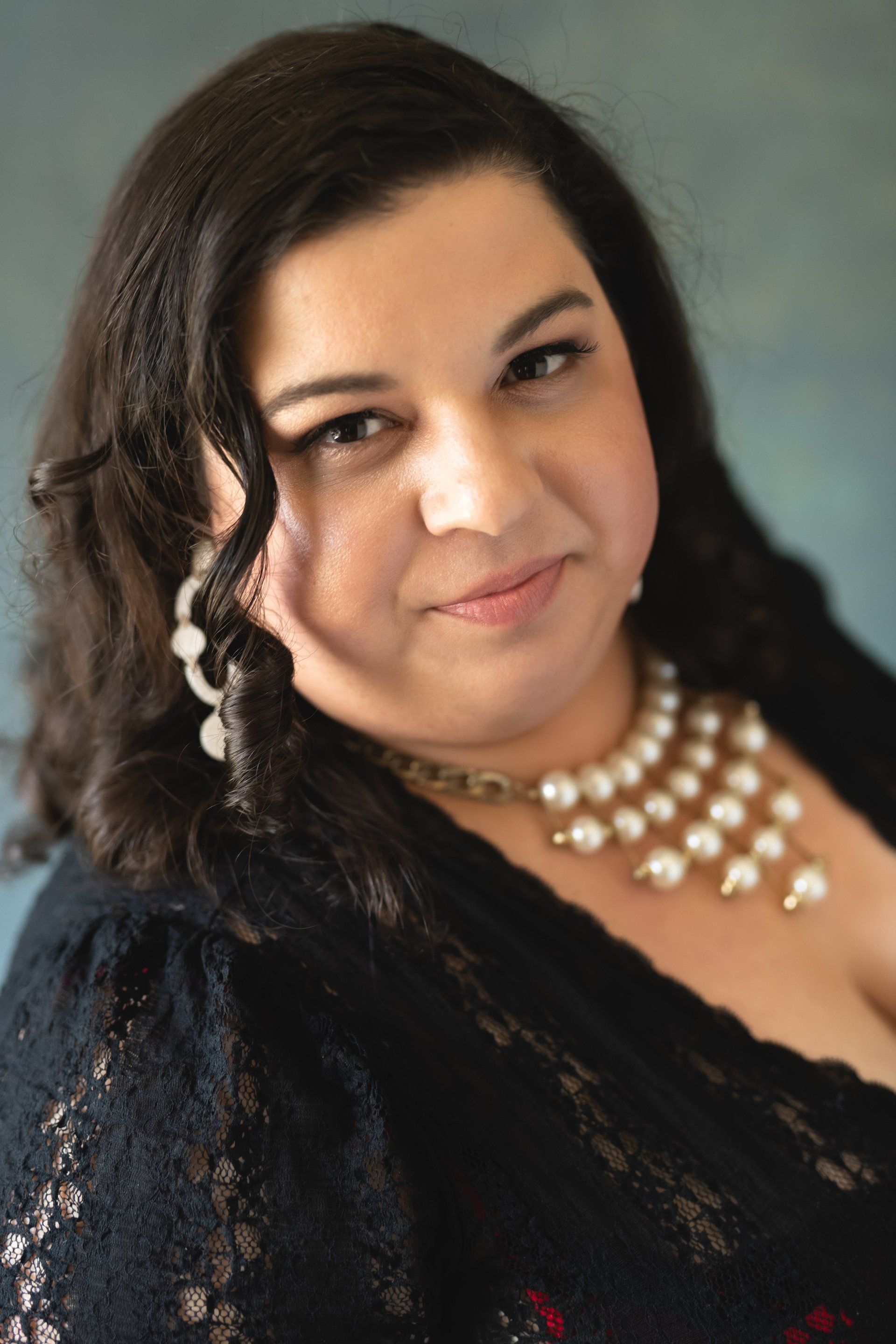 Woman with dark hair, wearing pearl necklace and black lace top, smiling.