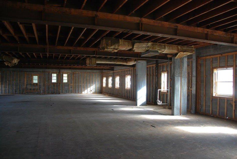 Empty unfinished room with exposed wooden framing, concrete floor, and sunlight through windows and doorways