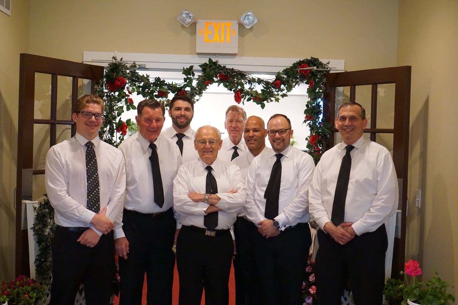 Seven people in white shirts and black ties pose indoors under a decorated archway.
