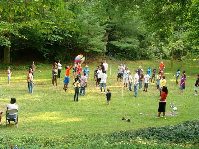 People gathered on a grassy field in a park, with trees in the background and a colorful umbrella near the center.