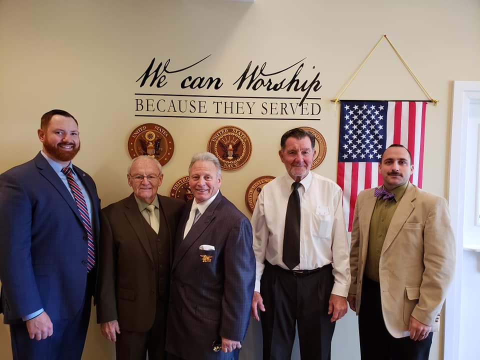 Five men in suits stand indoors beneath “We can worship because they served” wall sign and U.S. flag