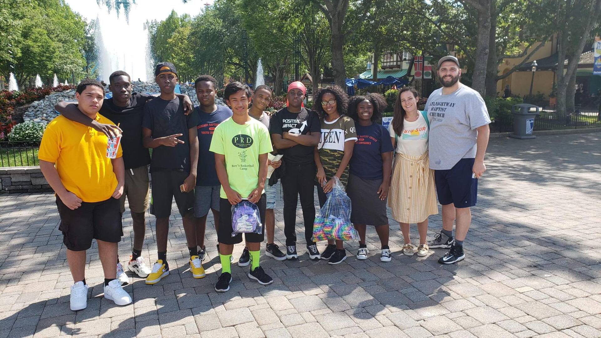 Group of people posing together outdoors on a sunny paved path near trees and fountains