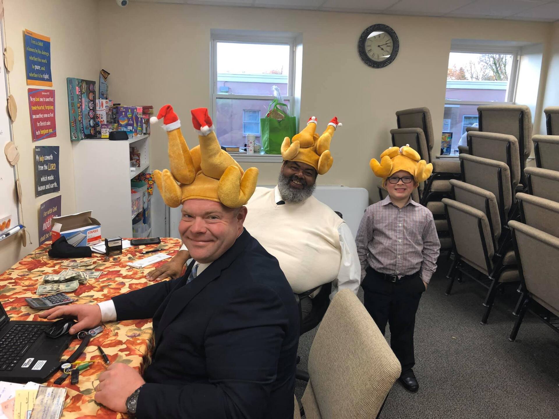 People wearing turkey hats sit and stand in a meeting room with tables, chairs, and windows.