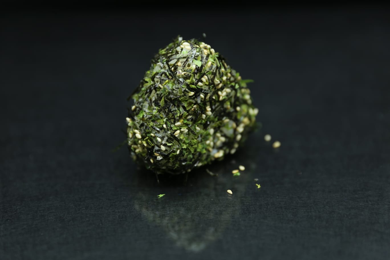 Un piccolo rotolo di sushi guarnito con scorza d'arancia e semi di sesamo nero, circondato da briciole d'arancia sparse su una superficie scura.