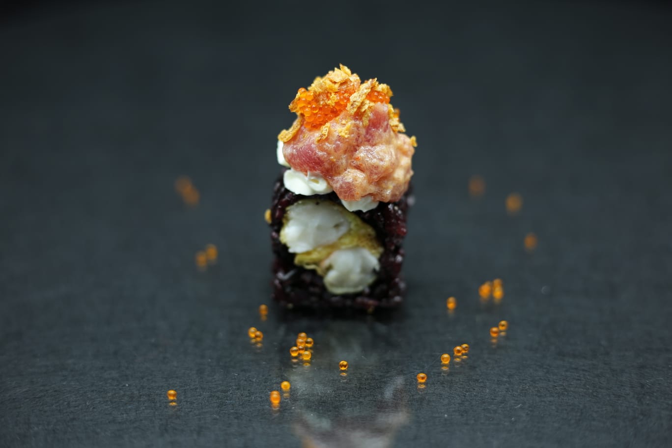 Un piccolo dessert a strati, guarnito con una decorazione arancione, al centro di una superficie scura e riflettente con briciole d'arancia sparse.