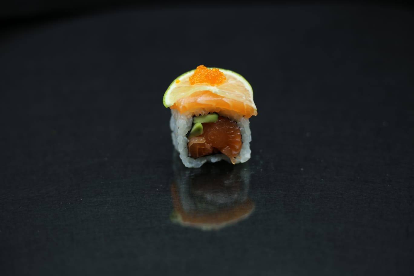 Pezzo singolo di sushi con guarnizione arancione, centrato su una superficie nera lucida con un debole riflesso