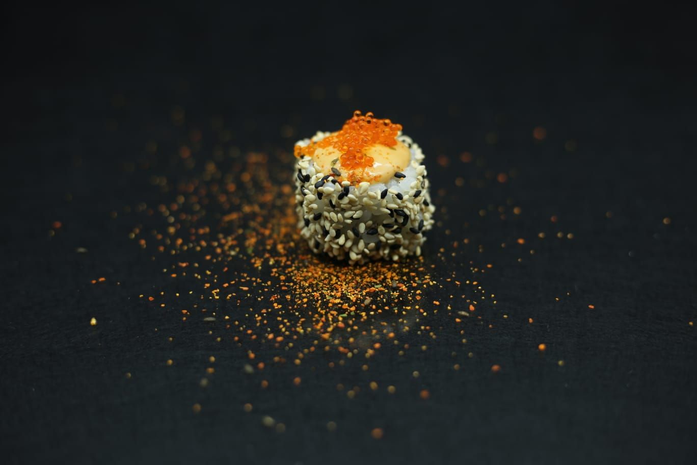 Rotolo di sushi singolo con guarnizione arancione su una superficie scura, cosparso di briciole di condimento.