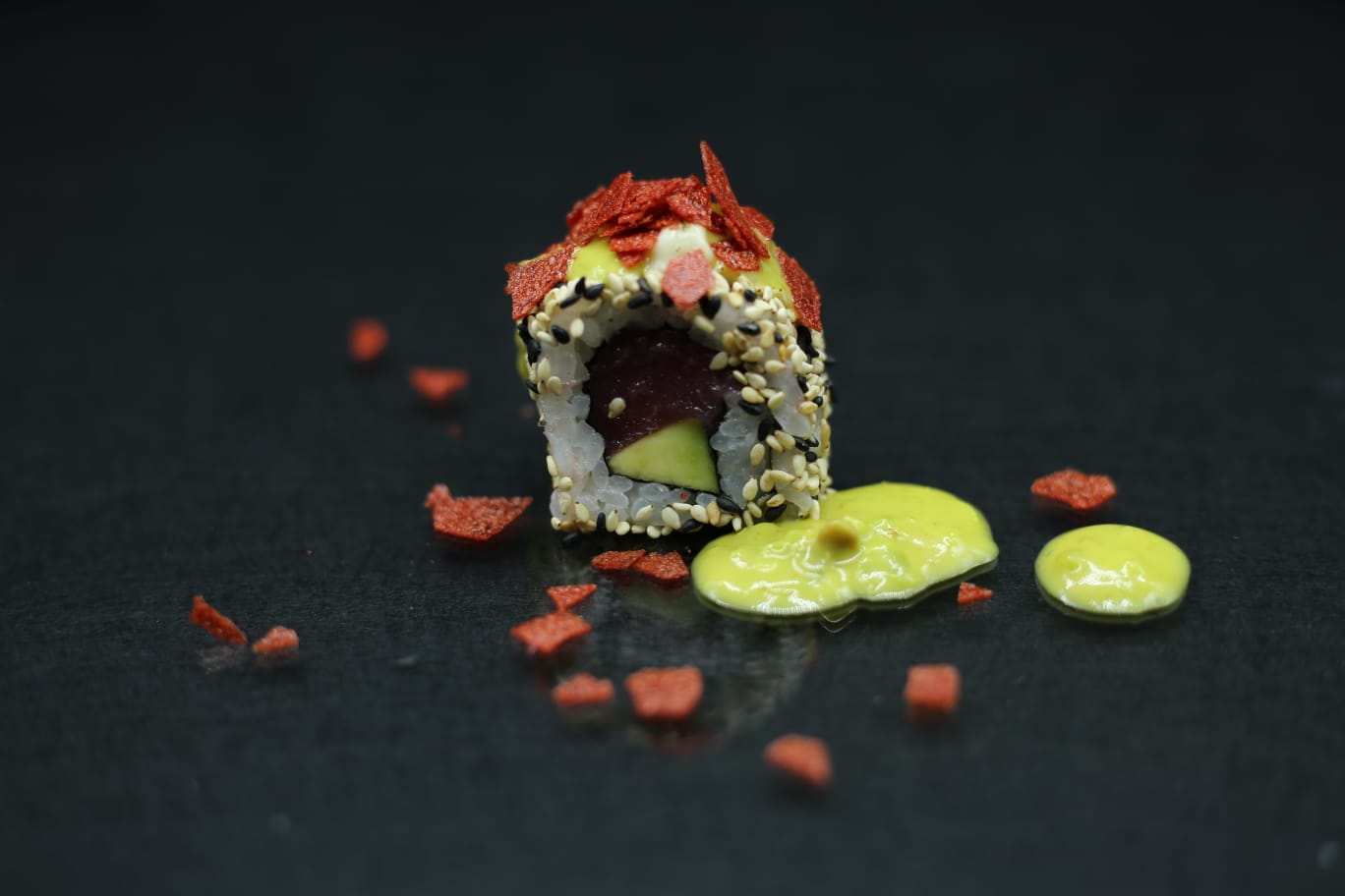Rotolo di sushi ricoperto di briciole rosse su una superficie scura, accanto a cucchiaiate di salsa verde
