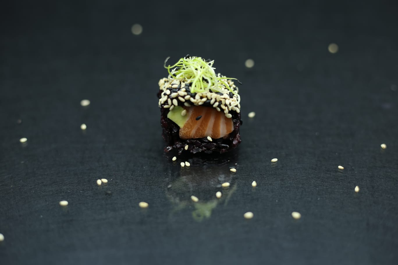 Piccolo rotolo di sushi guarnito con una decorazione verde e semi di sesamo su una superficie scura e riflettente.