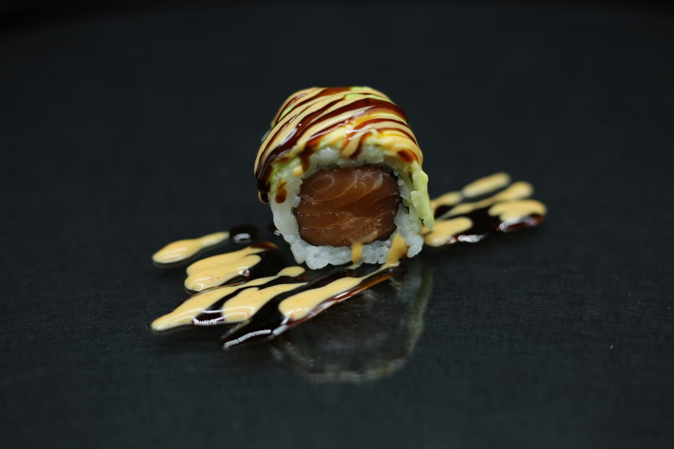 Sushi roll condito con salse scure e cremose, servito su un piatto nero.