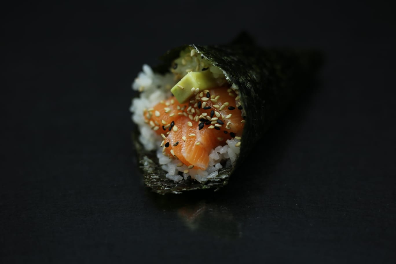 Primo piano di un rotolo di sushi con salmone, avocado e semi di sesamo su sfondo scuro.