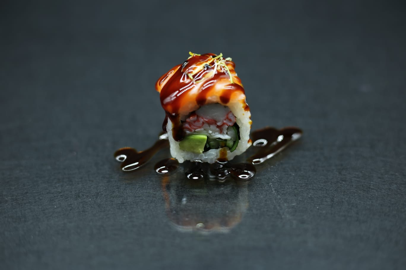 Rotolo di sushi ricoperto da una salsa lucida su una superficie scura e riflettente.