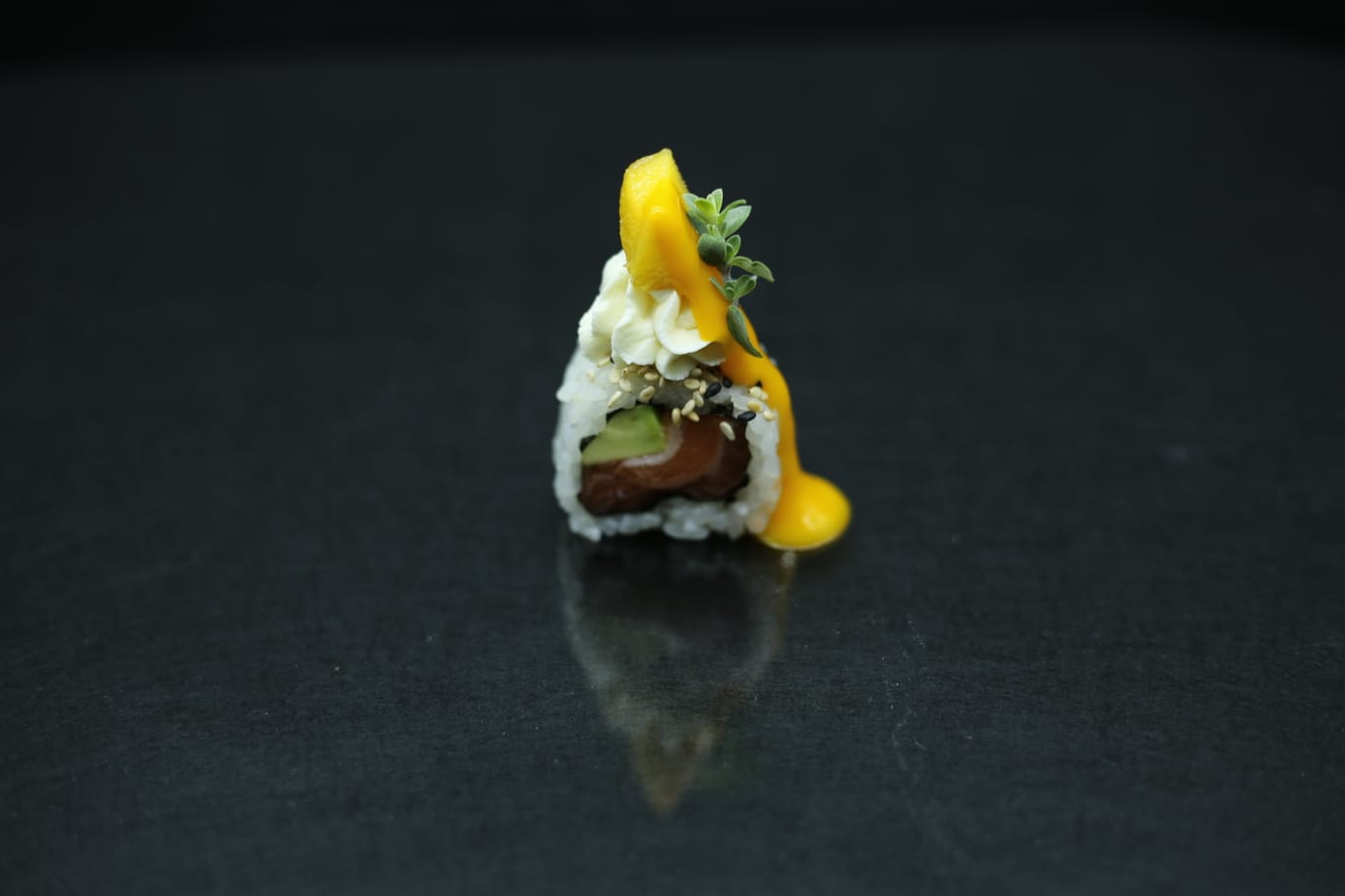 Un minuscolo rotolo di sushi ricoperto di salsa gialla su una superficie nera lucida.
