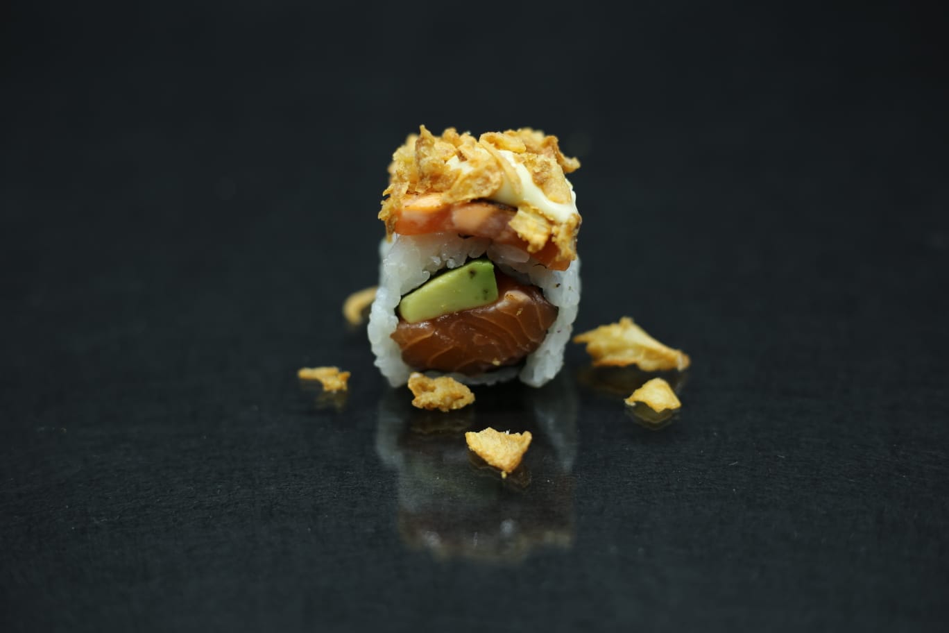 Primo piano di un rotolo di sushi con avocado su una superficie scura e riflettente, guarnito con scaglie croccanti.
