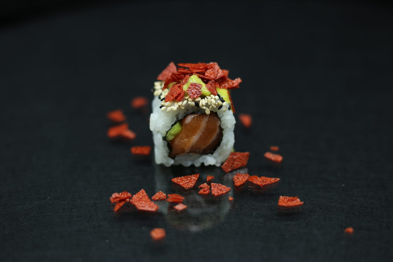 Primo piano di un rotolo di sushi condito con salsa ed erbe aromatiche, circondato da decorazioni rosse sparse su uno sfondo scuro.