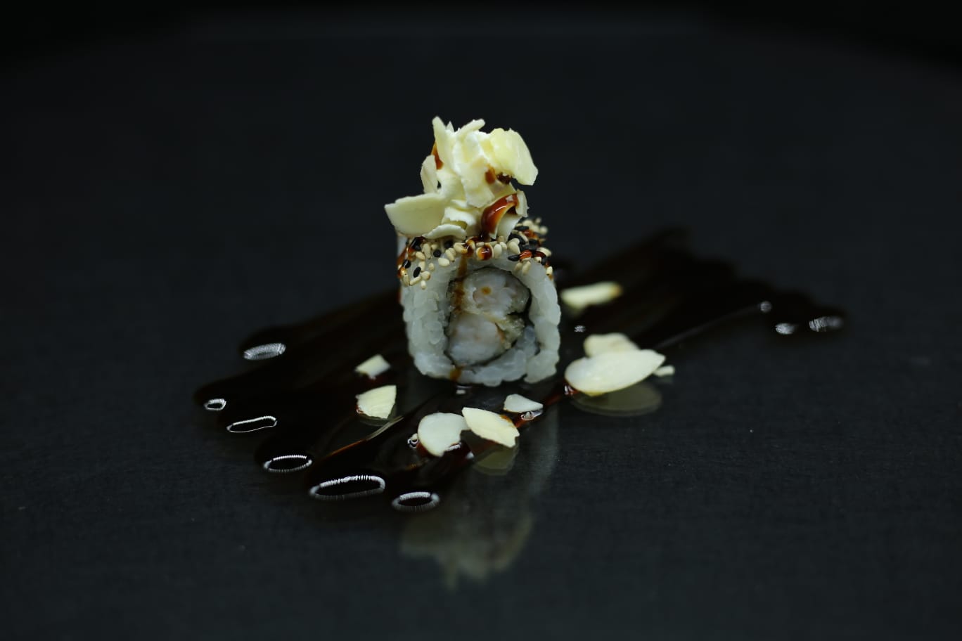 Primo piano di un rotolo di sushi con guarnizione bianca su un piatto nero, irrorato di salsa.