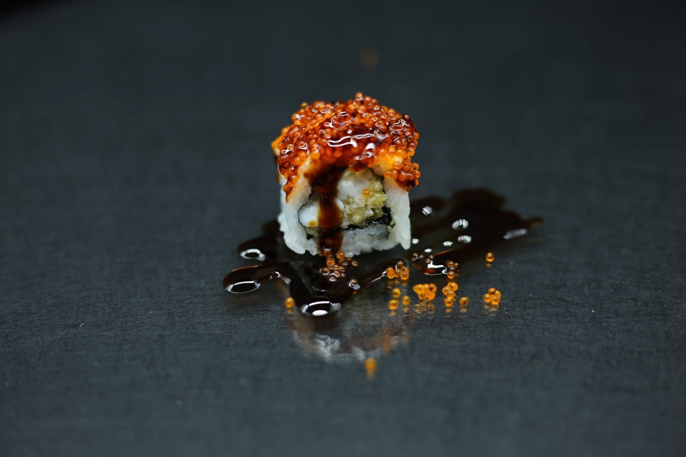 Piccolo rotolo di sushi con salsa scura e guarnizione arancione su una superficie nera.