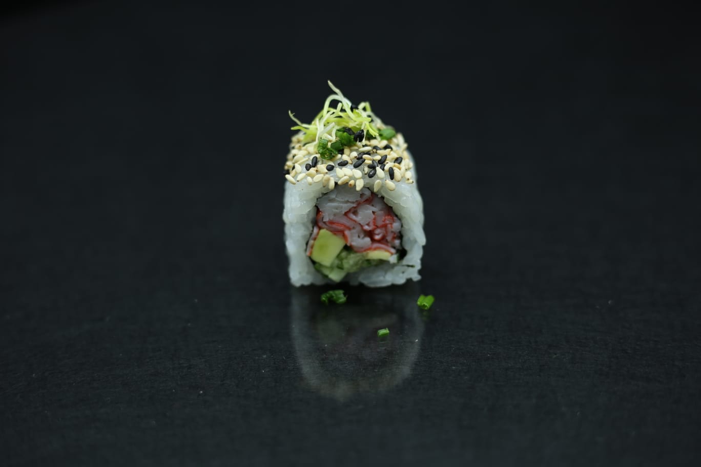 Rotolo di sushi su una superficie nera, guarnito con germogli e ripieno visibile al centro.