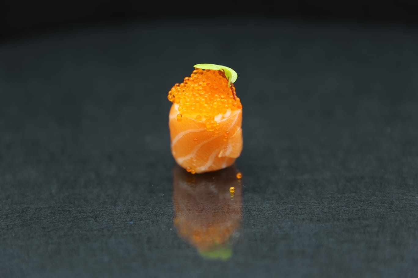 Un piccolo rotolo di sushi al salmone guarnito con uova di pesce arancioni su una superficie scura e riflettente.
